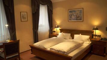 A hotel room with a double bed, two bedside tables with lamps, a desk with a chair, a window with curtains, and a painting on the wall.