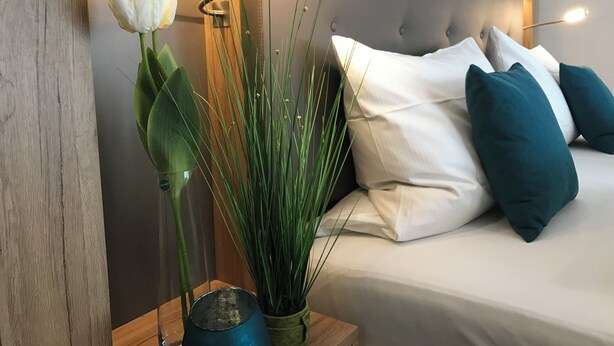 A bed with white and teal pillows, a bedside table with a vase of white flowers, and a tall green plant. - Hotel Kronprinz (Kulmbach)