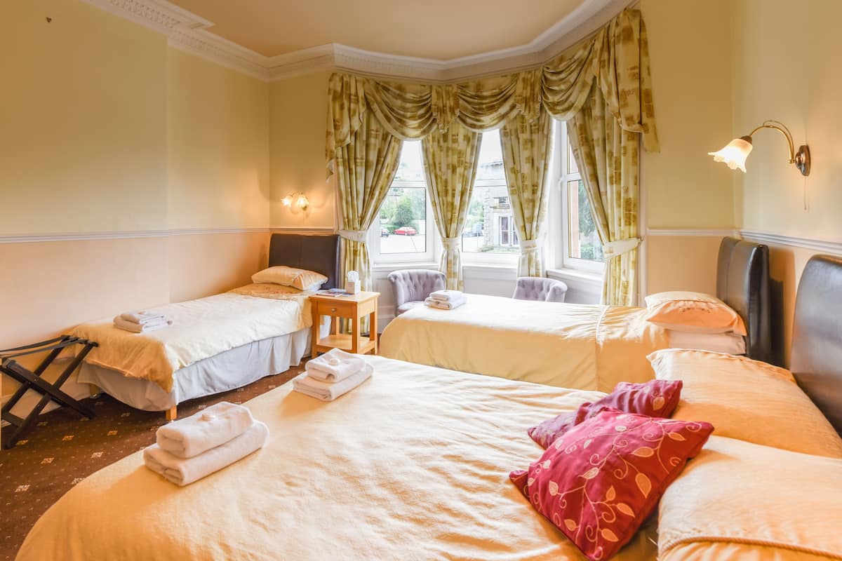 A hotel room with two single beds, a large bed, and a chair. There are towels on the beds and a small table with a lamp.
