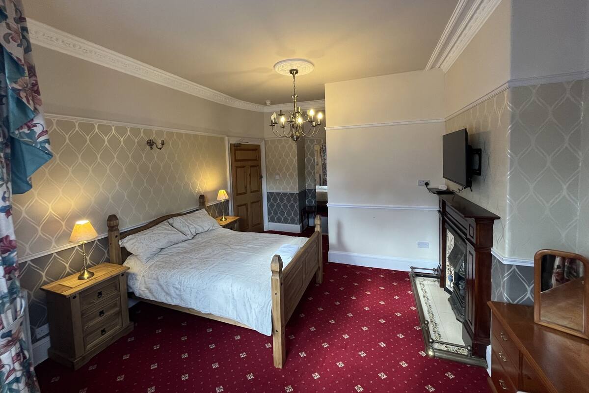 A bedroom with a bed, bedside tables, a chandelier, a television, and a fireplace.
