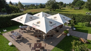 An outdoor patio with multiple tables and chairs, covered by large umbrellas with the brand 'Benediktiner'. - Venngasthof Zur Buche (Monschau)