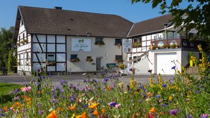 A traditional half-timbered building with a sign indicating it is a restaurant. There are bicycles parked in front, and a variety of flowers in the foreground. - Venngasthof Zur Buche (Monschau)