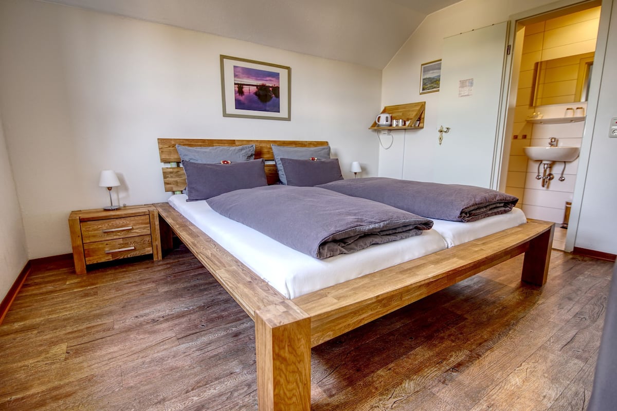 Una cama doble con estructura de madera y ropa de cama gris, una mesita de noche con lámpara y suelo de madera.