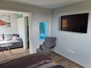 Een moderne woonkamer met een grijze bank, een klein rond tafeltje, een grijze fauteuil, een bed met donker beddengoed en een flatscreen televisie aan de muur.