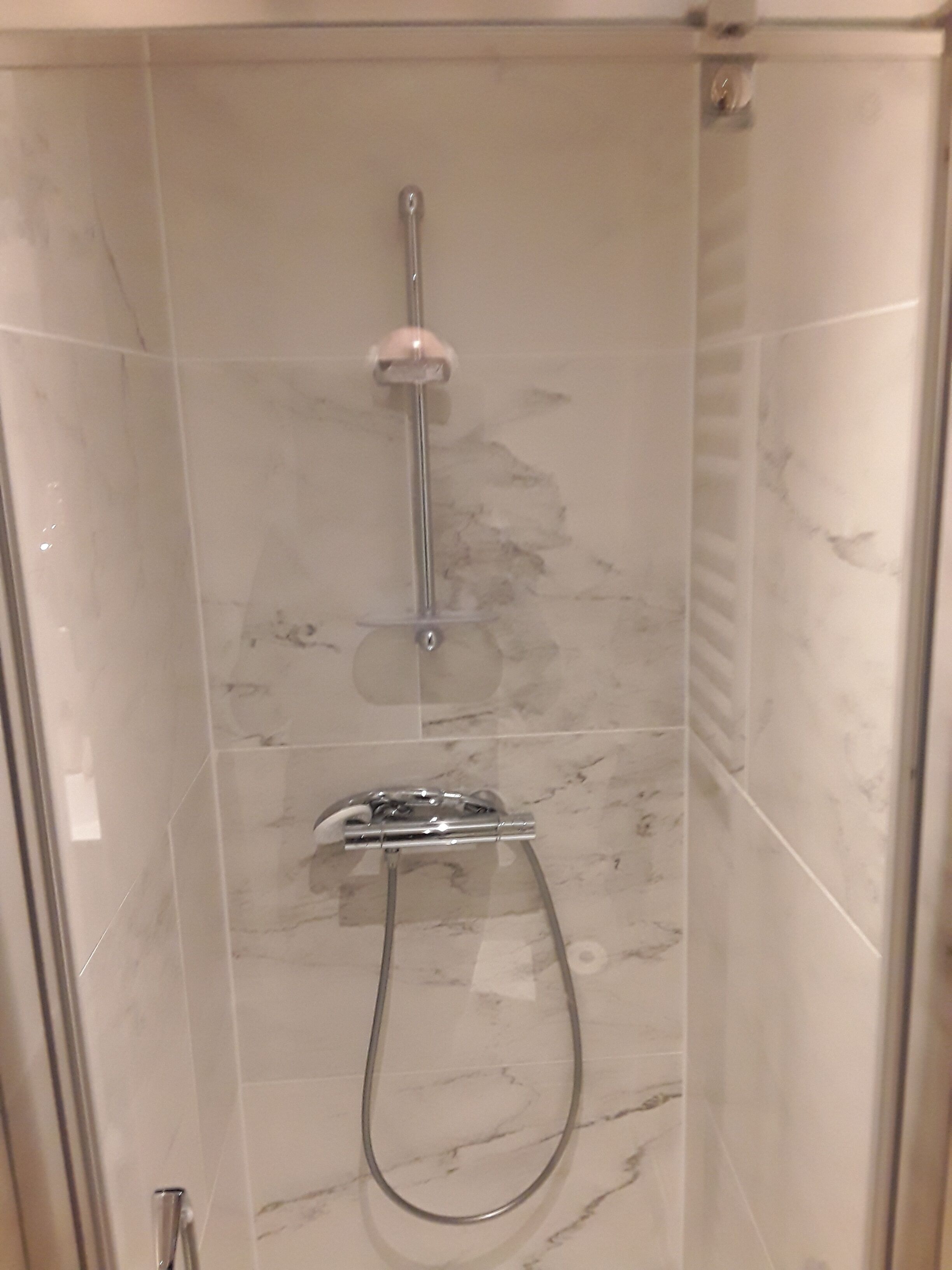 Une douche équipée d’un pommeau de douche à main et d’un pommeau de douche fixe au plafond, tous deux fixés sur un mur recouvert de carreaux imitant le marbre.