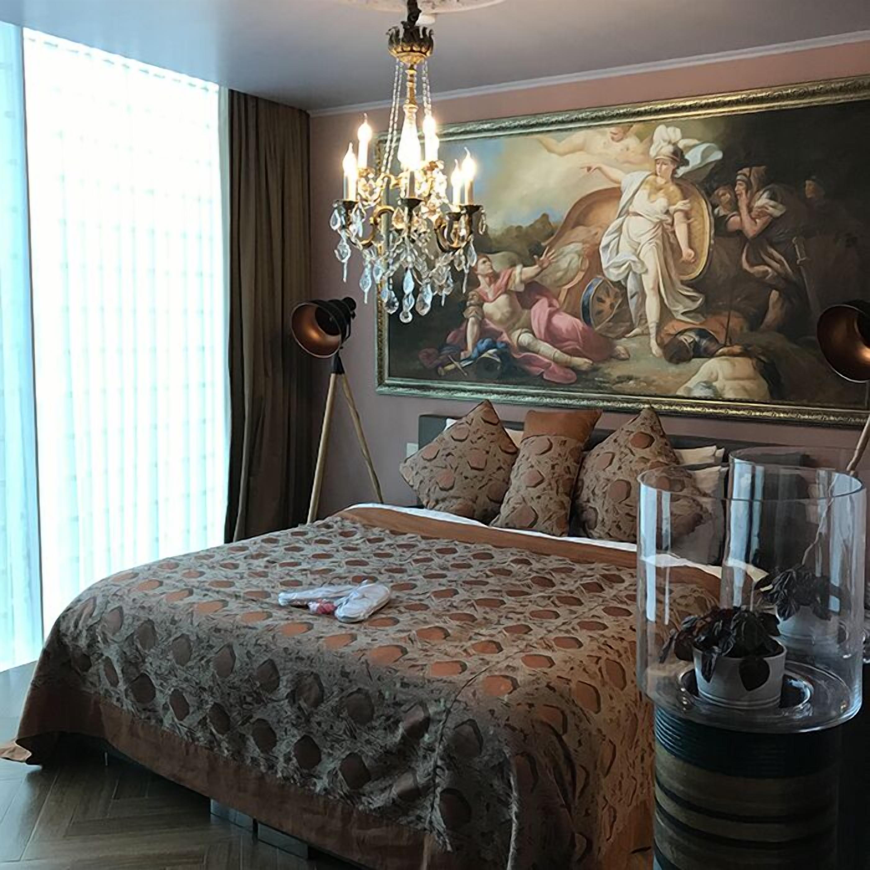 A bedroom with a large bed, a chandelier, a painting, and a floor lamp.