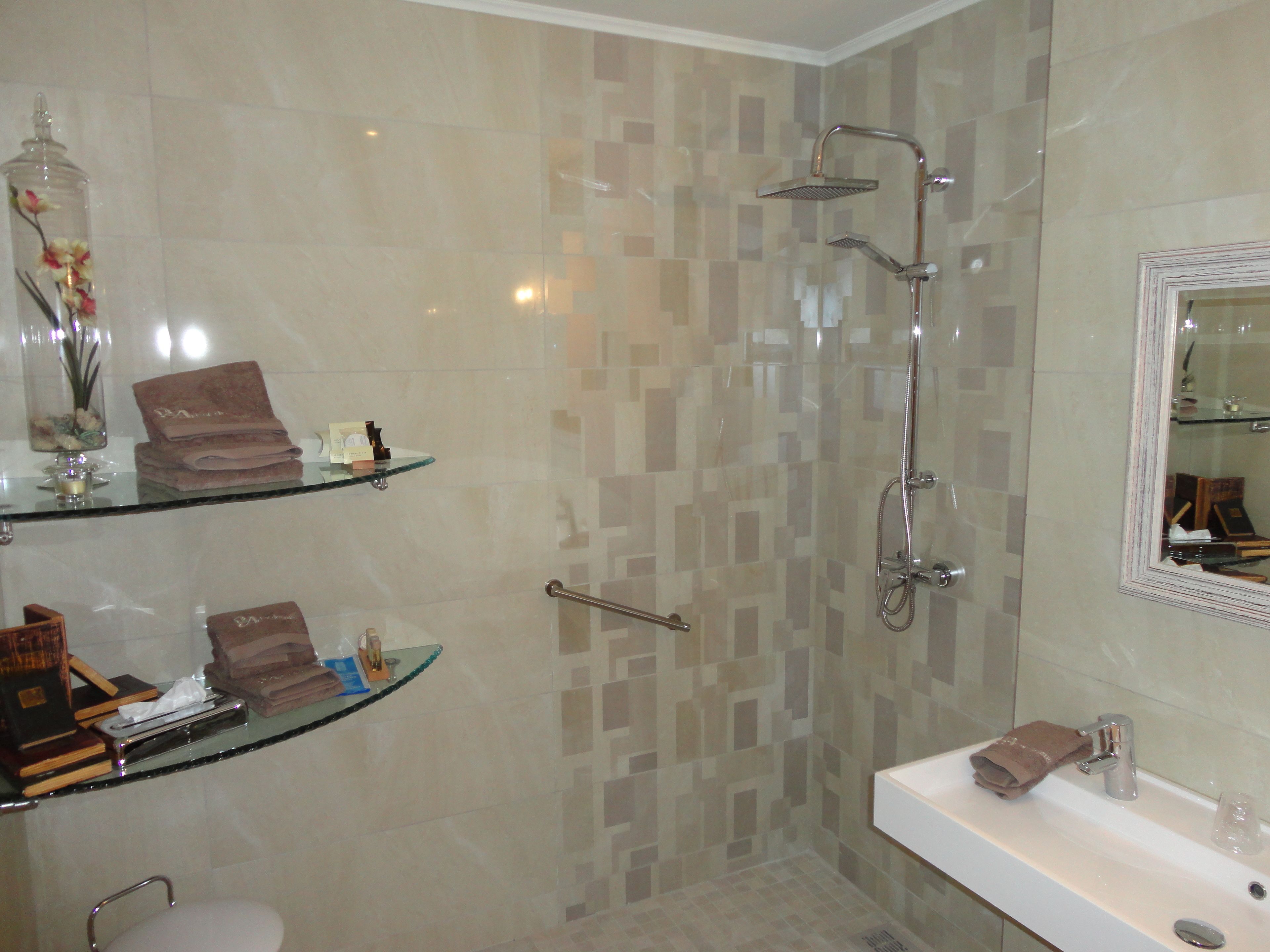 A bathroom with a glass shelf holding toiletries, a shower with a handheld showerhead, and a vanity with a mirror.