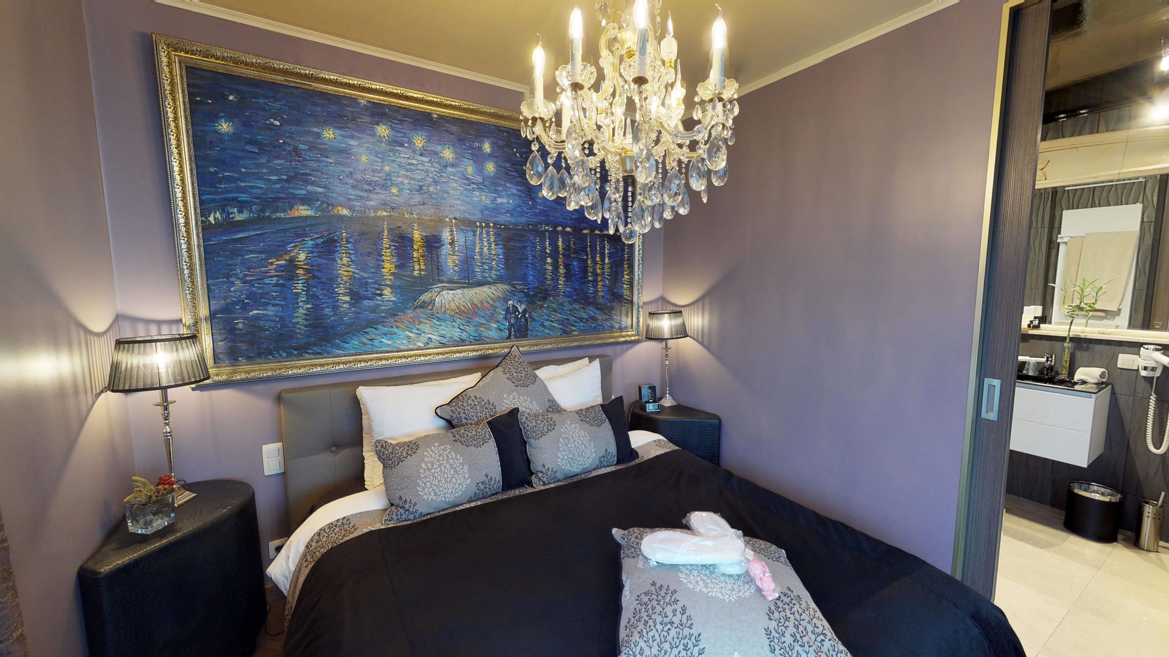 Een slaapkamer met een groot schilderij van een nachtelijke scène, een kroonluchter en een moderne badkamer die door een open deur zichtbaar is.
