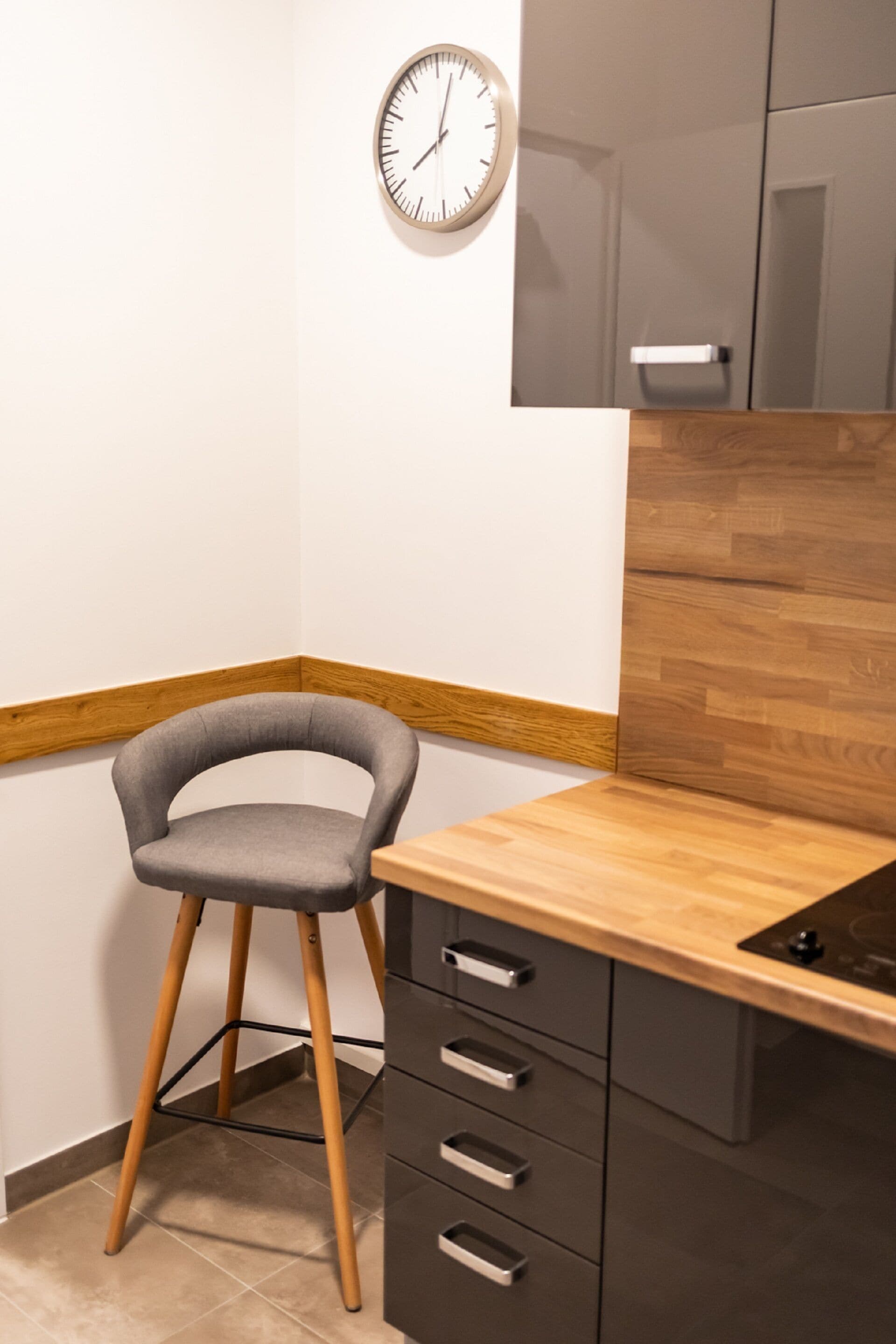 Un rincón de una habitación con un escritorio de madera, una silla gris, un reloj de pared y un armario.