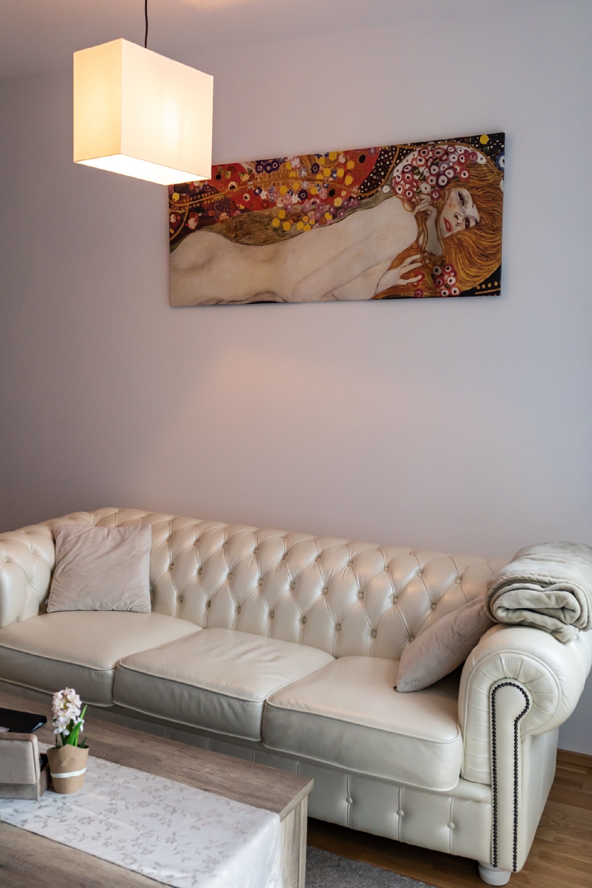 Un sofá color beige con textura, un cojín decorativo, una mesita con un florero y una obra de arte colgada en la pared.