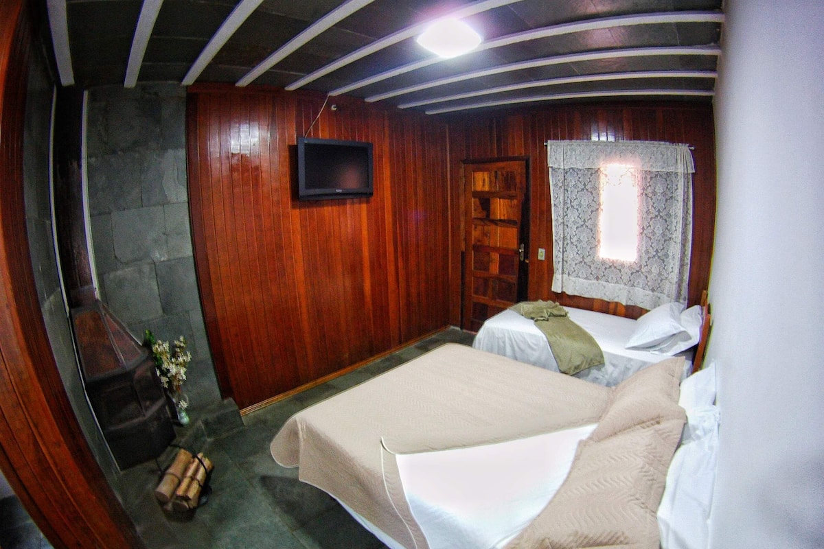 A room with wooden walls, a stone floor, a bed with white bedding, a small table with a vase, and a television on the wall.