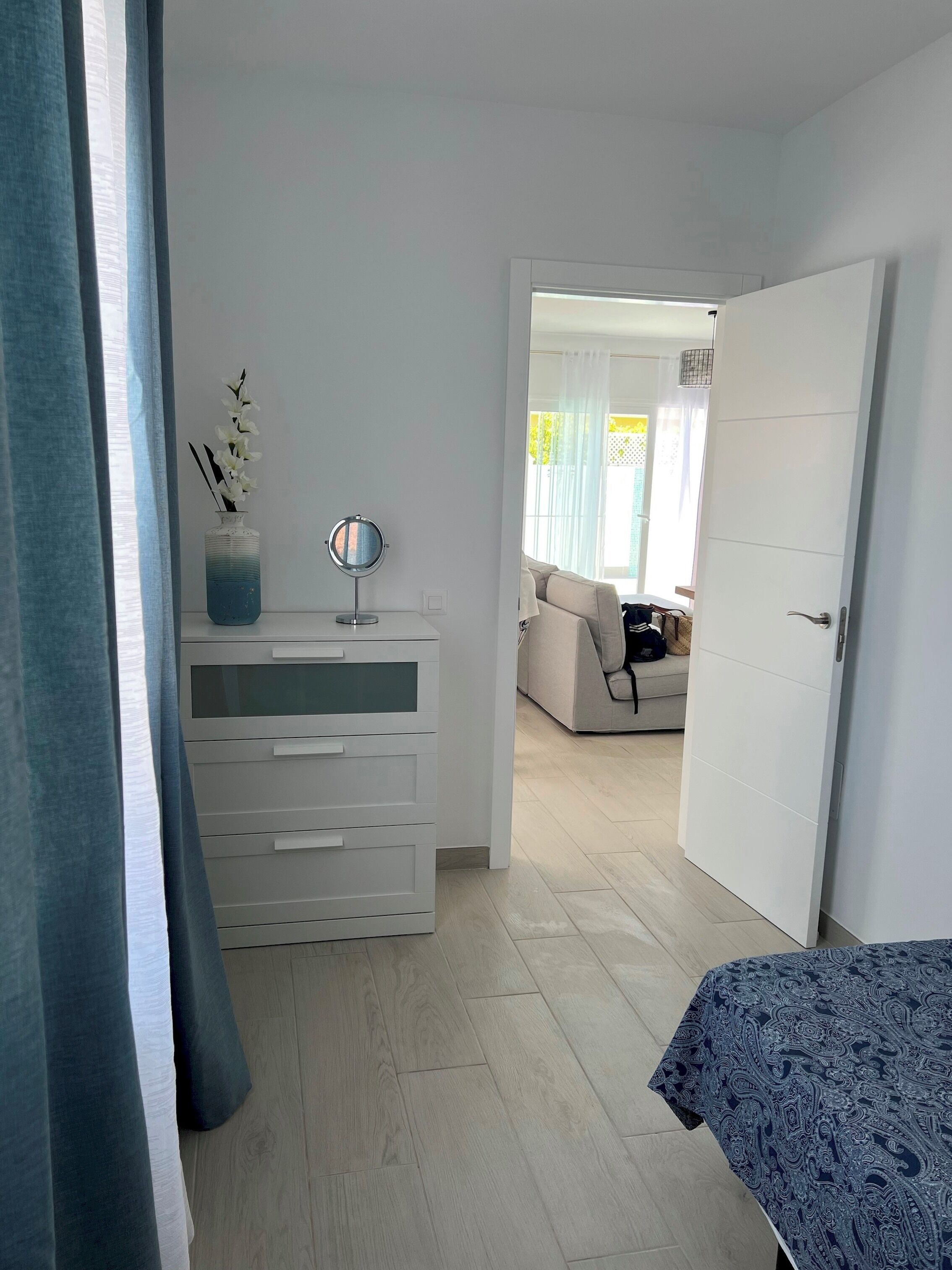 Un dormitorio con cama, cómoda, espejo y un florero con flores. Se ve una puerta que da a otra habitación.