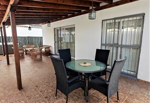 Une terrasse couverte avec une table ronde et des chaises, un plafond en bois et une vue à travers une fenêtre grillagée.