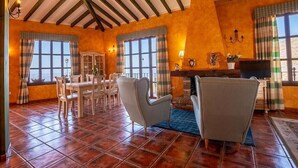 Il s’agit d’une pièce spacieuse avec un plafond haut, un sol carrelé et de grandes fenêtres dotées de rideaux. On y trouve une cheminée, une table à manger avec des chaises et un canapé.