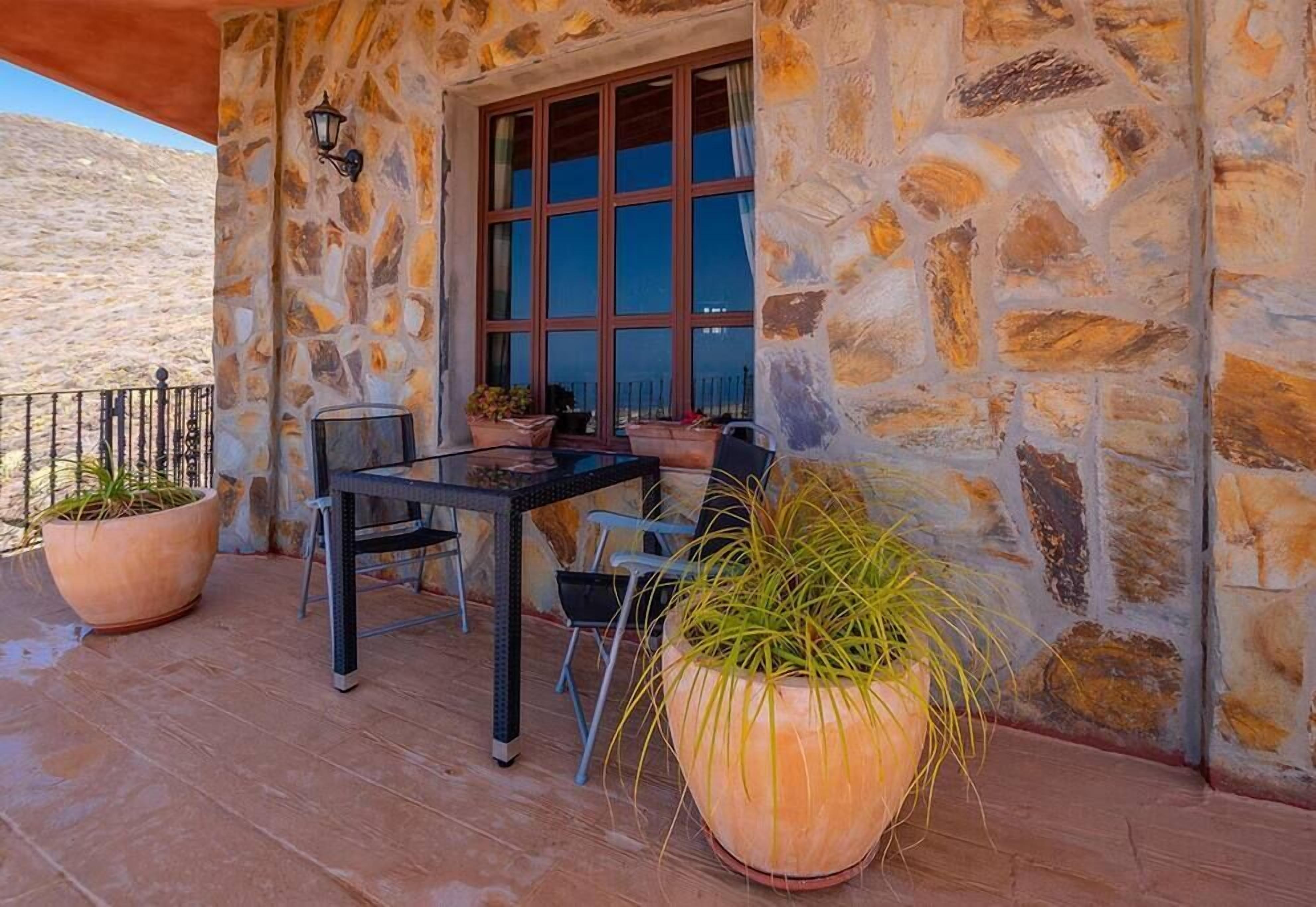 A stone-walled outdoor area with a table and chairs, potted plants, and a window with multiple panes.