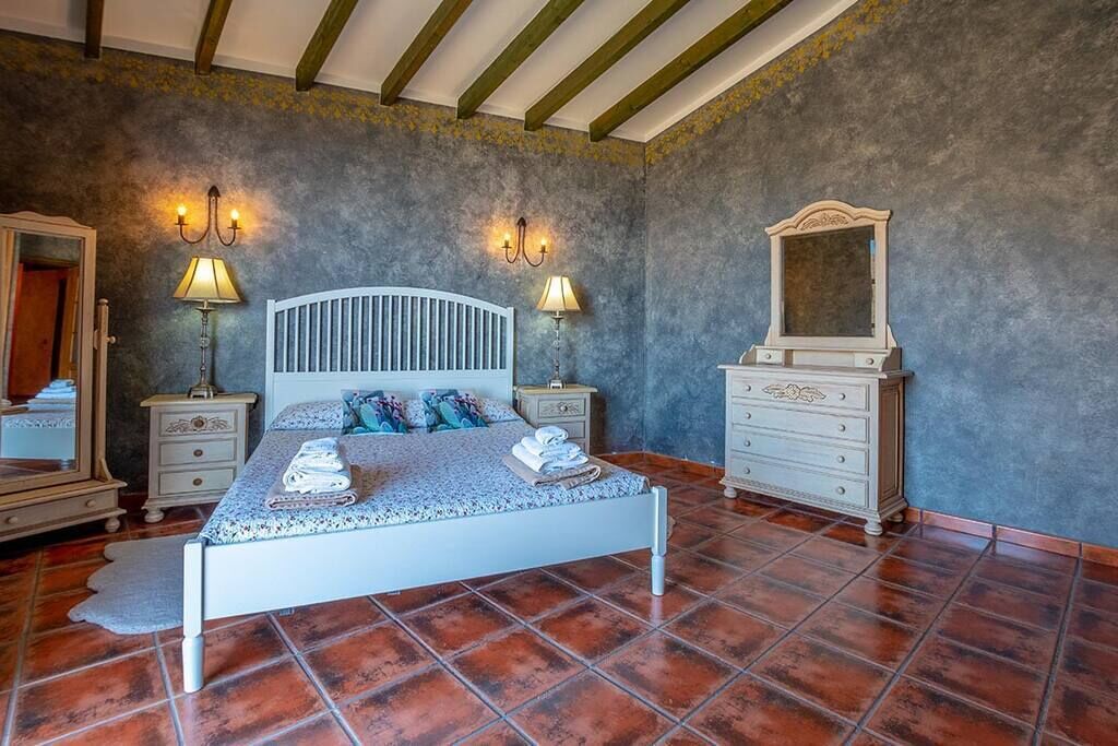 A bedroom with a bed, two bedside tables, a dresser, a mirror, and wall sconces.