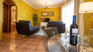 Un salon aux murs jaunes, avec un lustre, un canapé, un téléviseur et une bouteille de vin posée sur une table.
