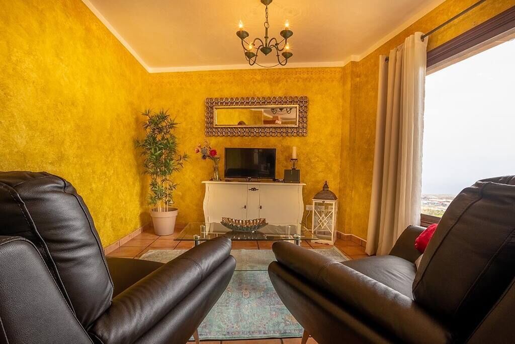 A living room with yellow walls, a white cabinet, a TV, a chandelier, and a view through a window.