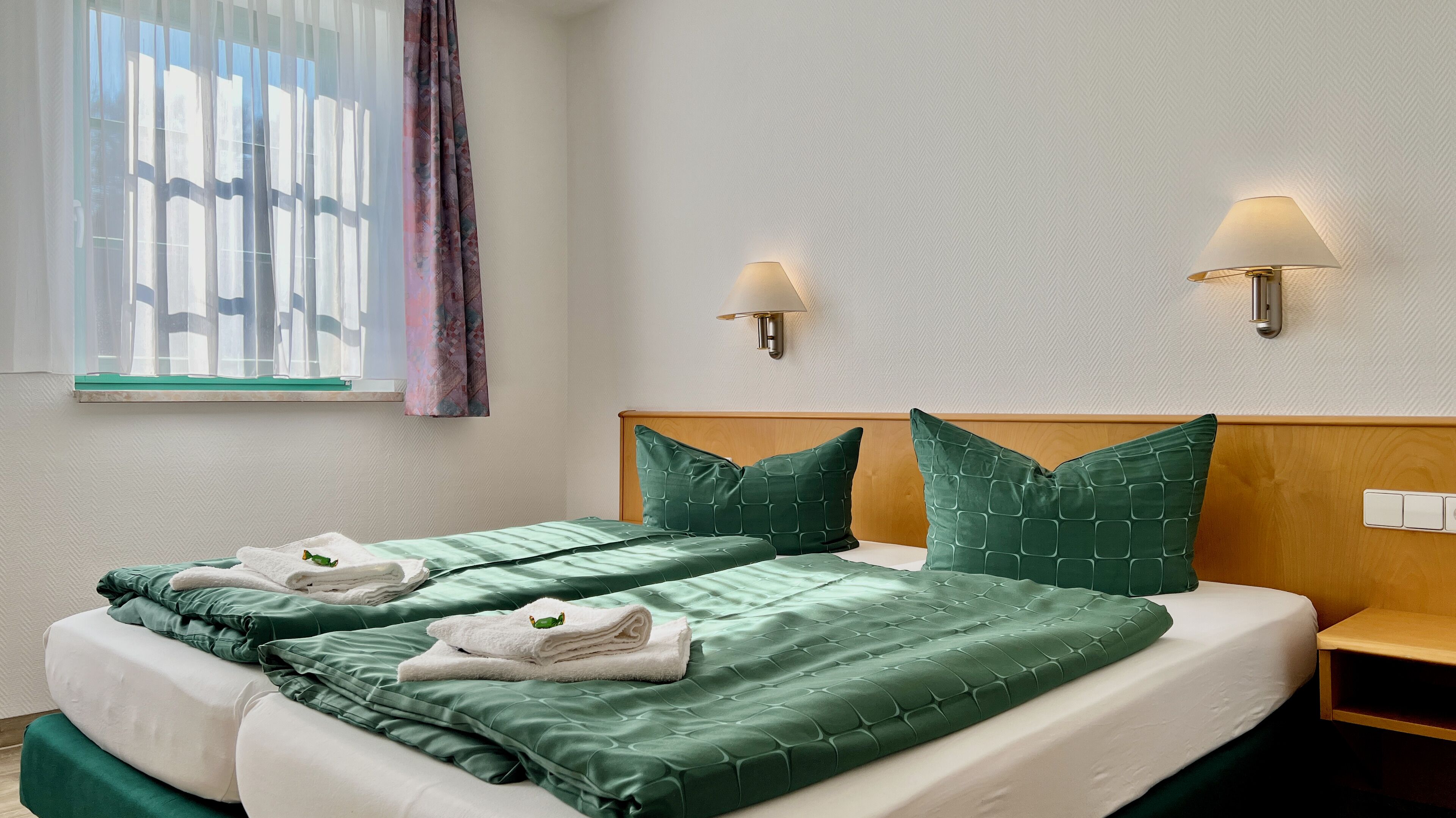 A hotel room with two beds, green bedding, a wooden headboard, two wall-mounted lamps, and a window with sheer curtains.