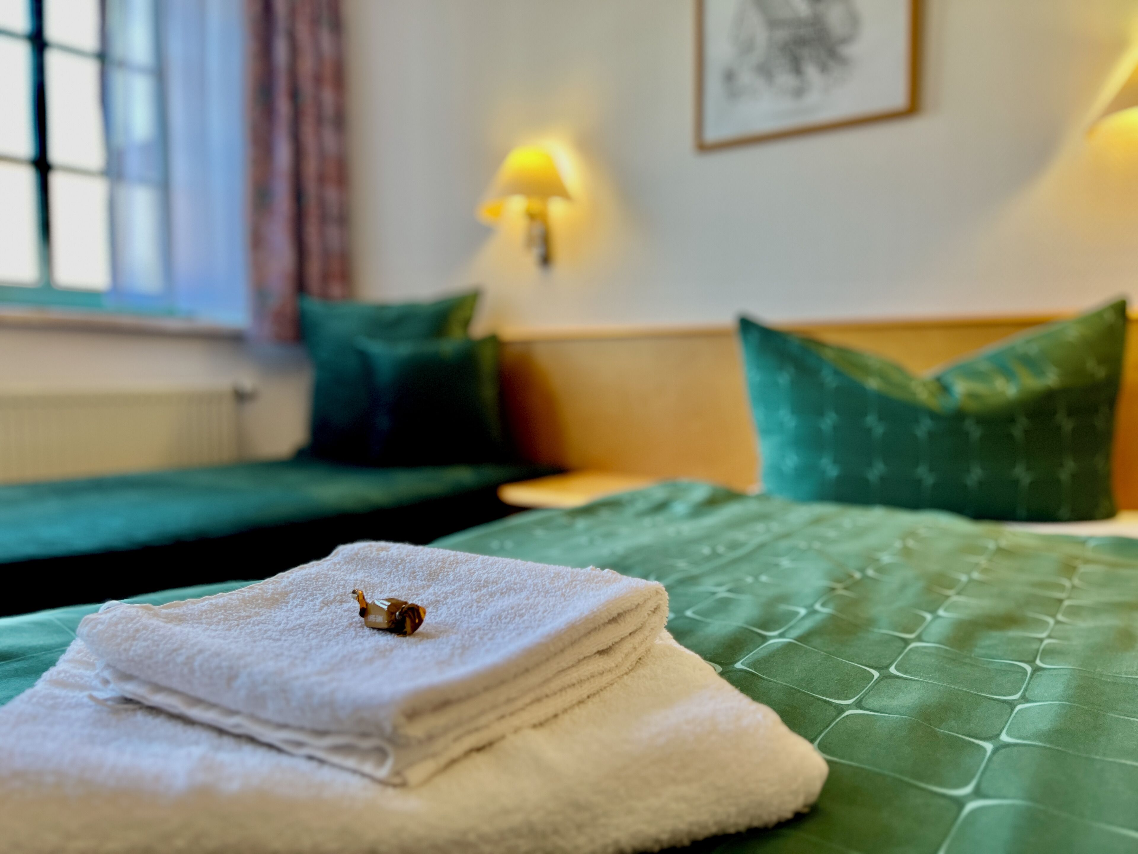 A neatly made bed with green bedding and a folded white towel with a small decorative item on top.