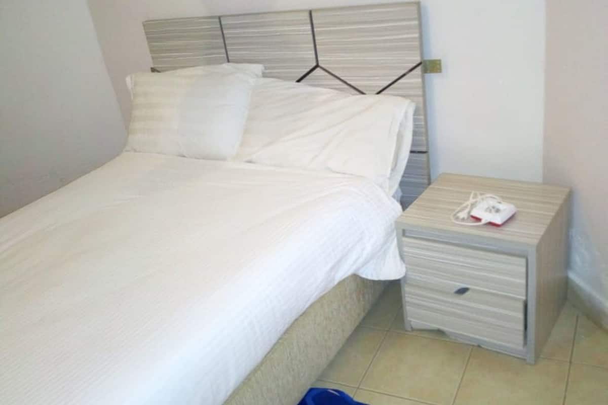 A neatly made bed with white linens, a bedside table with a phone and charger, and a blue bag on the floor.