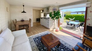 Amplia sala de estar con sofá, una mesa de centro y un mueble para televisión. Hay un comedor con mesa y sillas, y una cocina con armarios blancos y un microondas. La habitación tiene un gran ventanal corredizo que da a un patio exterior con mesa y sillas.