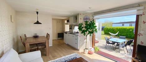 A spacious living room with a sofa, a coffee table, and a TV stand. There is a dining area with a table and chairs, and a kitchen with white cabinets and a microwave. The room has a large sliding glass door leading to an outdoor patio with a table and chairs.