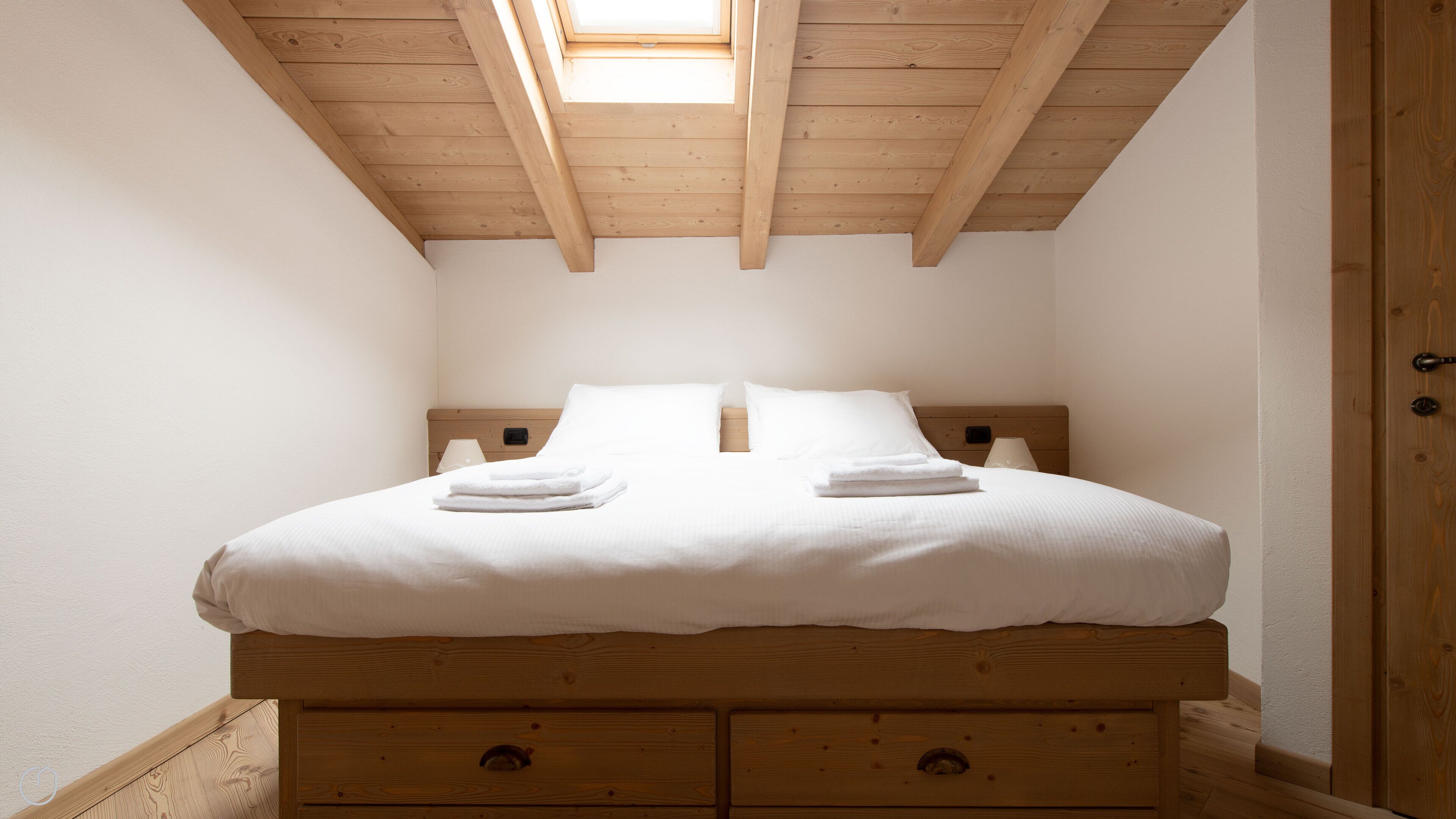 Una cama individual con cabecera de madera, dos mesitas de noche con lámparas y un tragaluz superior.