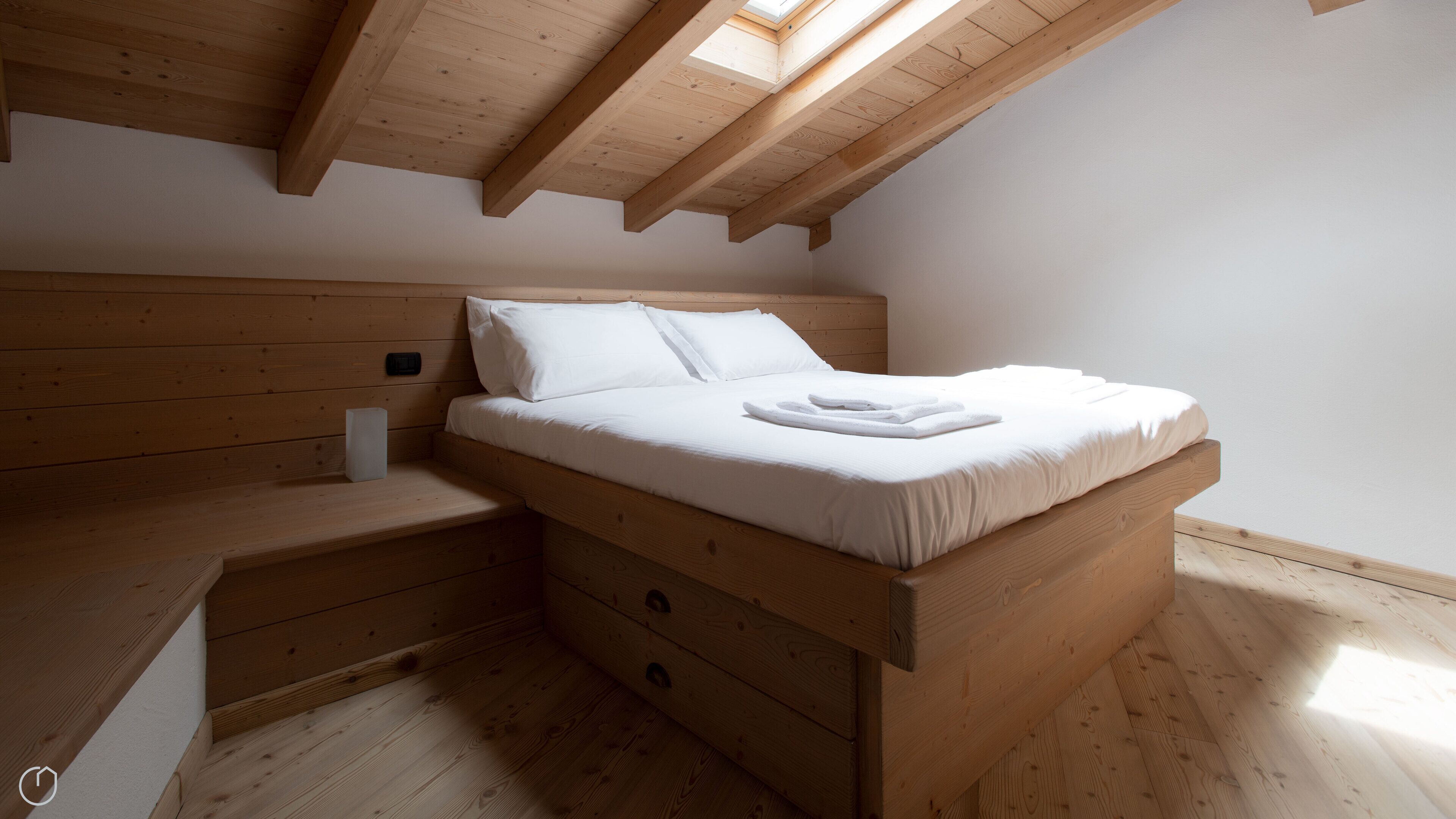 A wooden loft bedroom with a sloped ceiling, a bed with white bedding, and a small bedside table.