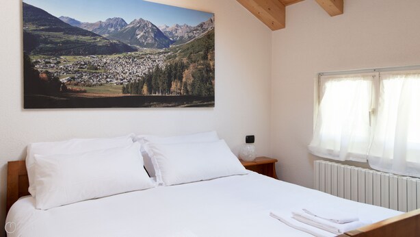 Une chambre à coucher avec un grand lit, du linge de lit blanc, une table de chevet avec une lampe, une fenêtre avec des rideaux légers et un tableau représentant un paysage de montagne accroché au mur.
