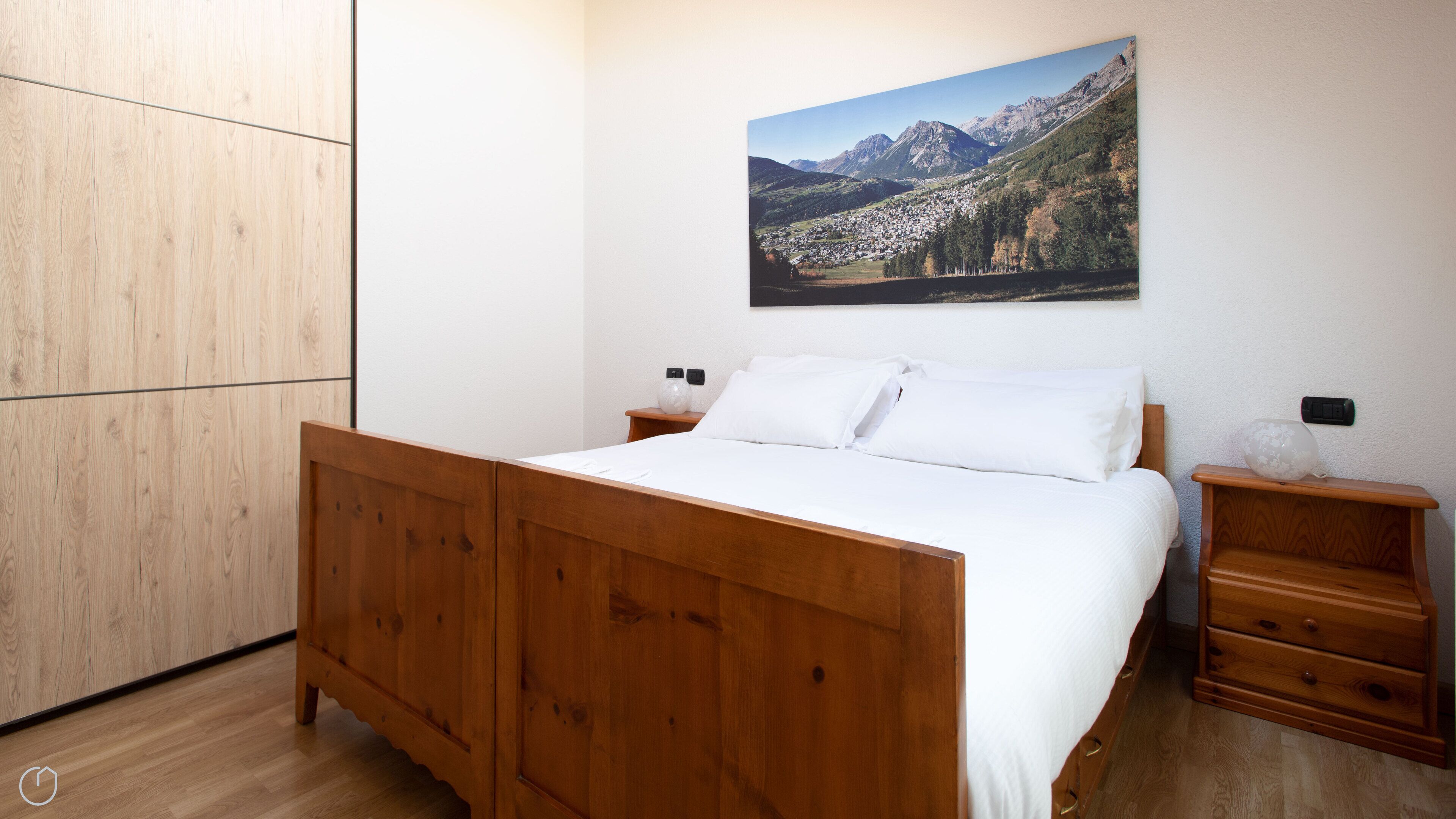 A bedroom with a wooden bed, white bedding, a nightstand with a vase, and a painting of a mountain landscape.