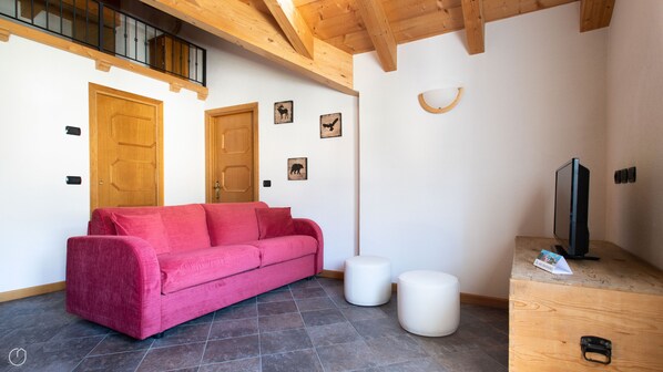Quarto com sofá vermelho, dois taburetes brancos, televisão em suporte de madeira e teto de madeira com varanda.