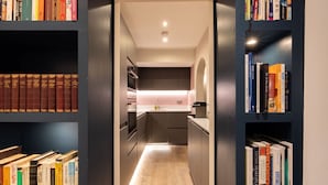 A narrow hallway with a kitchen area, bookshelves on either side, and a wooden floor. - The Brunswick Oasis 2 bedroom in Notting Hill (London)