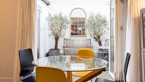 A glass table with chairs, a bench, and two potted plants in a room with white walls and a window. - The Brunswick Oasis 2 bedroom in Notting Hill (London)
