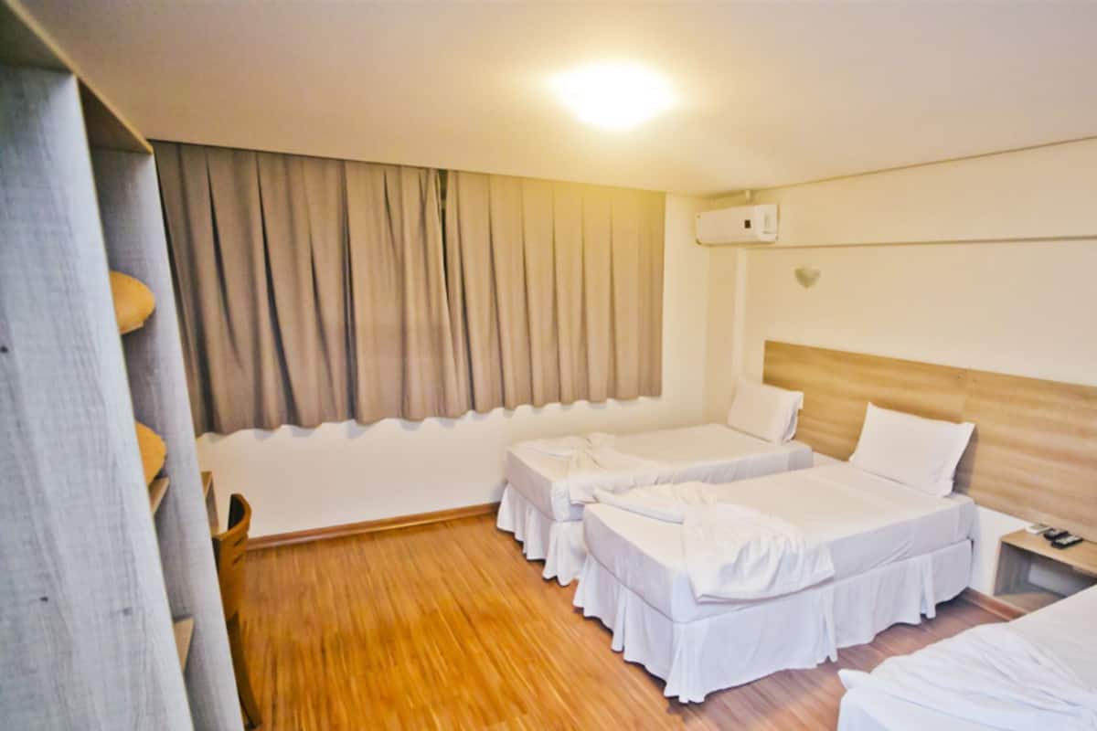 A hotel room with two single beds, a wooden floor, a large window with curtains, and a bedside table with a lamp.