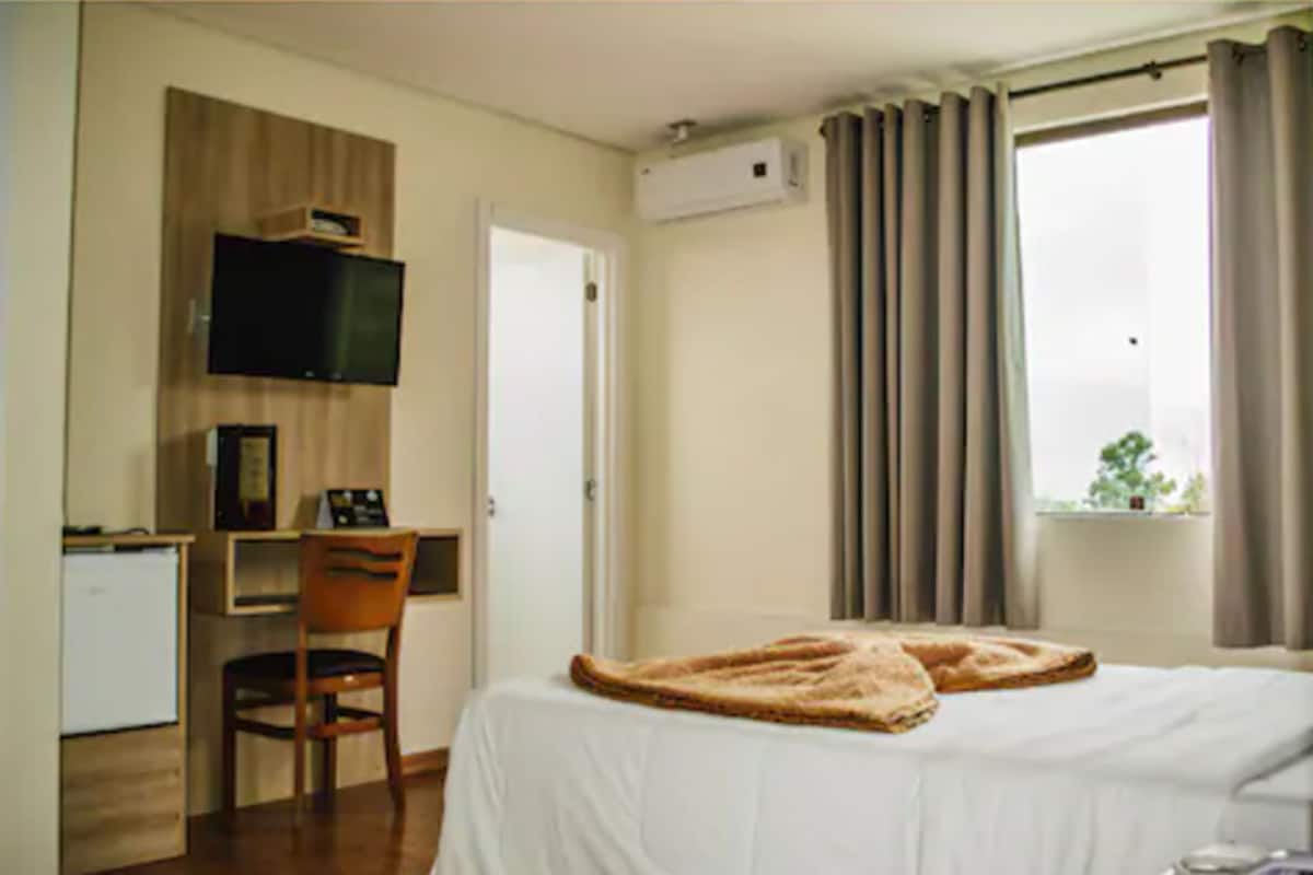 A hotel room with a bed, a desk with a chair, a television mounted on the wall, and a window with curtains.