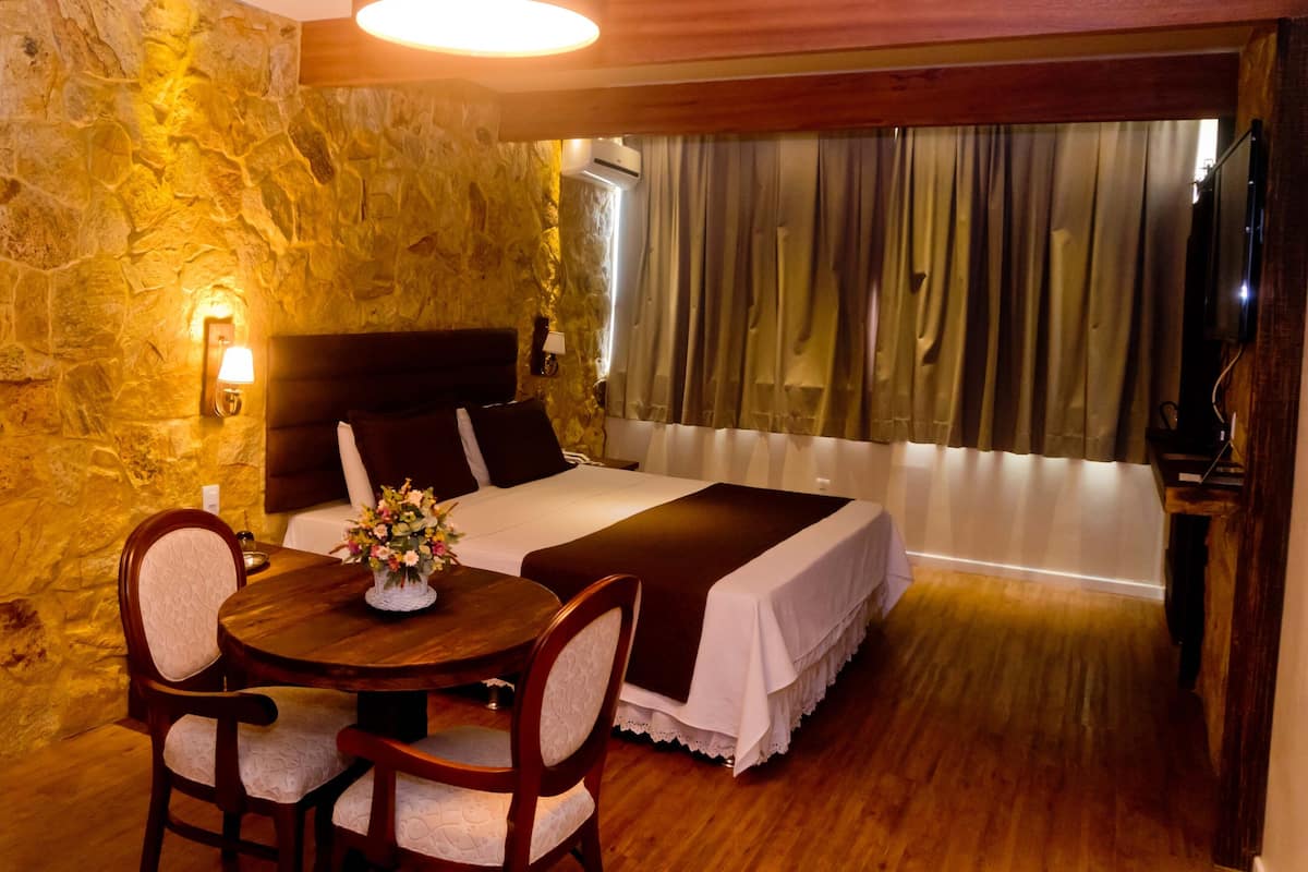 A hotel room with a stone wall, a bed with a headboard, a dining table with chairs, and a window with curtains.
