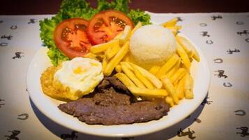 A plate with a beef patty, French fries, a fried egg, a tomato slice, and a white round item, possibly a potato or bread, on a table with a patterned tablecloth.