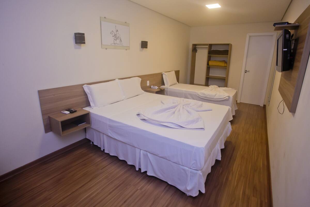 A hotel room with two beds, a wooden headboard, a bedside table, a wall-mounted TV, and a small shelf.