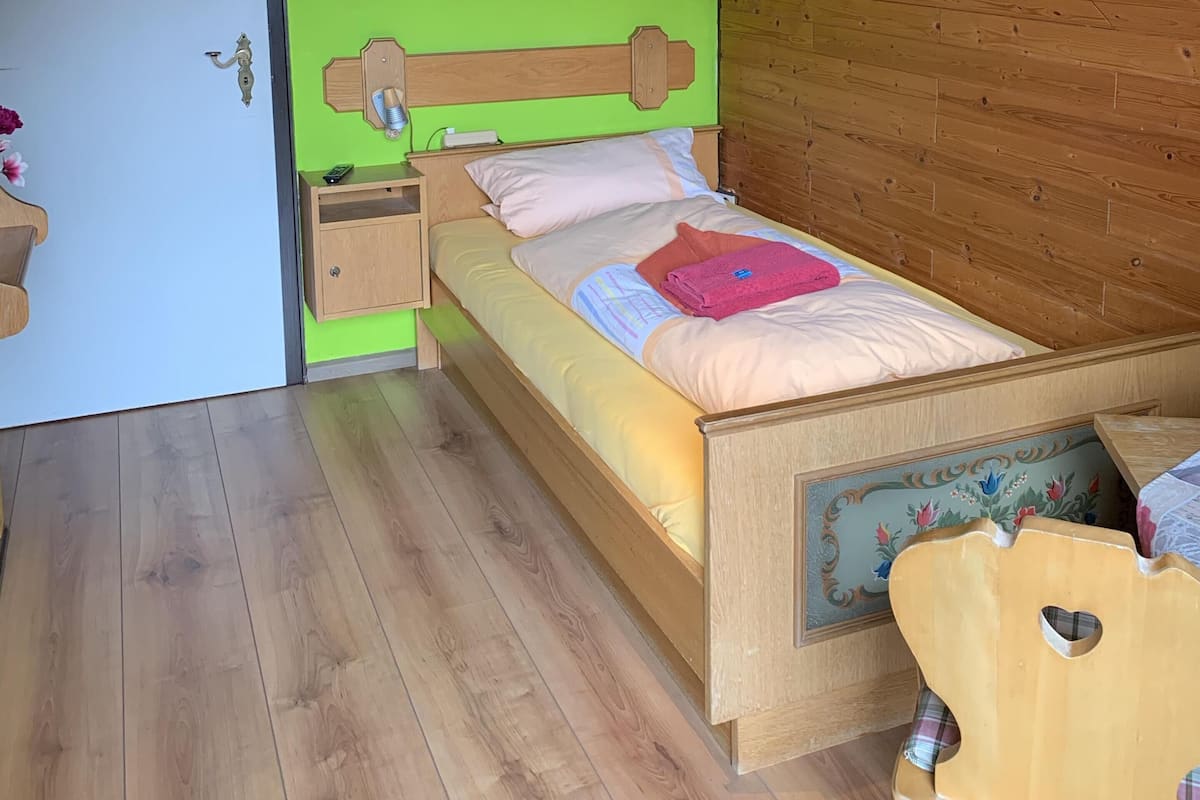 A room with a sloped ceiling, wooden walls, a bed with a wooden headboard, a bedside table, a nightstand, and a small wooden chair.