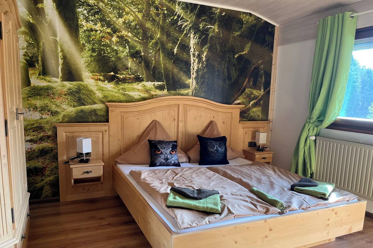A bedroom with a large bed, two nightstands, a wall mural, and a window with curtains.