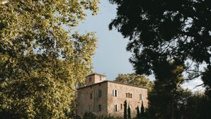 Un edificio de piedra con múltiples ventanas y una pequeña torre, rodeado de árboles y un muro de piedra.