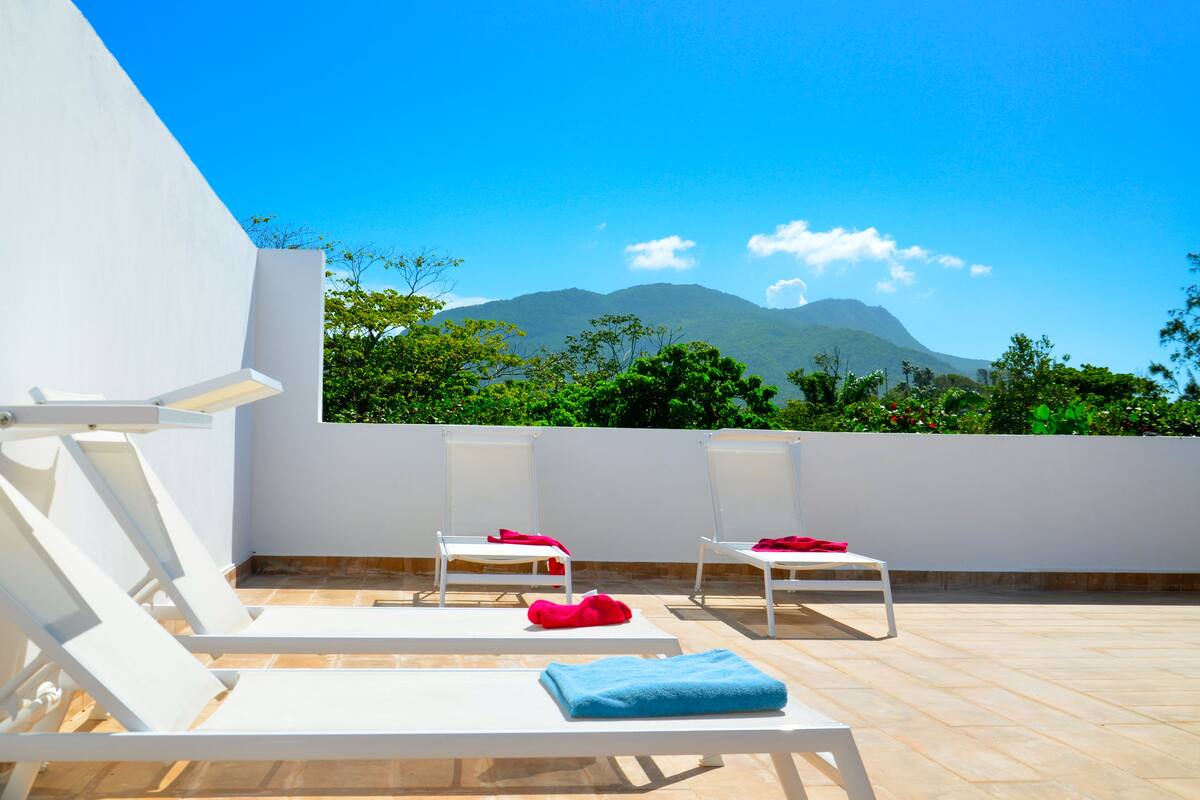 Une terrasse sur le toit, dotée de chaises longues blanches, dont certaines sont agrémentées de serviettes rouges et bleues, et offrant une vue sur des arbres verdoyants et des montagnes.