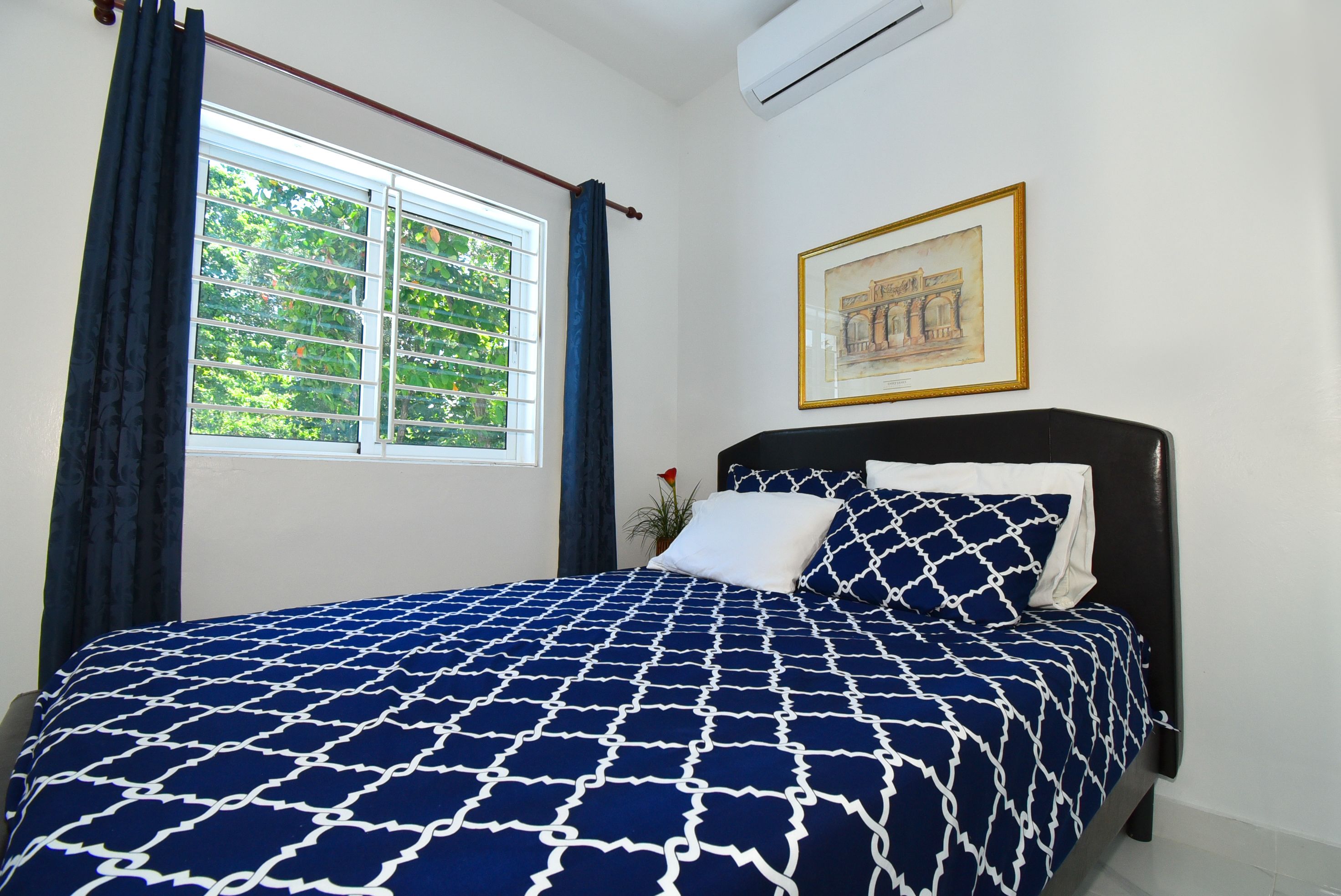 Ein Schlafzimmer mit einem Bett, einem Fenster mit Vorhängen, einem gerahmten Bild an der Wand und einer Klimaanlage.