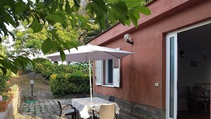En uteplats med ett bord och stolar under ett vitt parasoll, en röd byggnadsvägg och en dörr som leder in till ett inre utrymme.