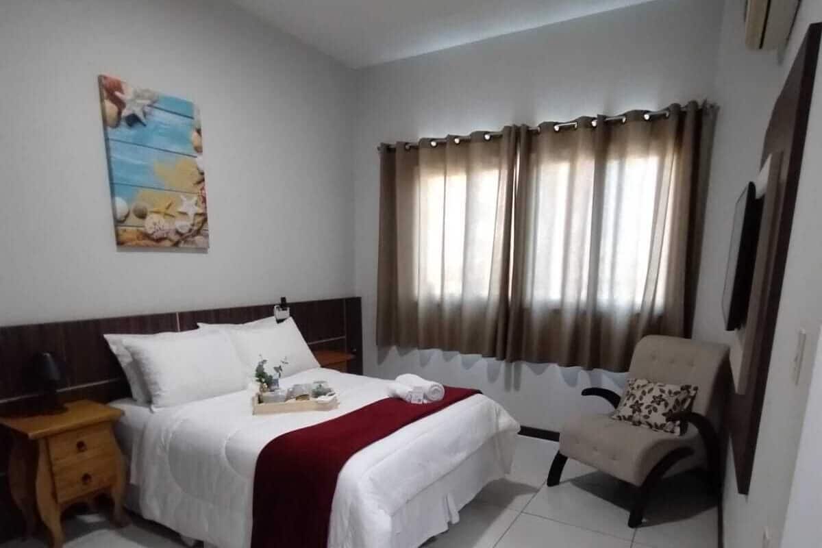 A hotel room with a bed, a chair, a nightstand, a painting on the wall, and curtains on the window.