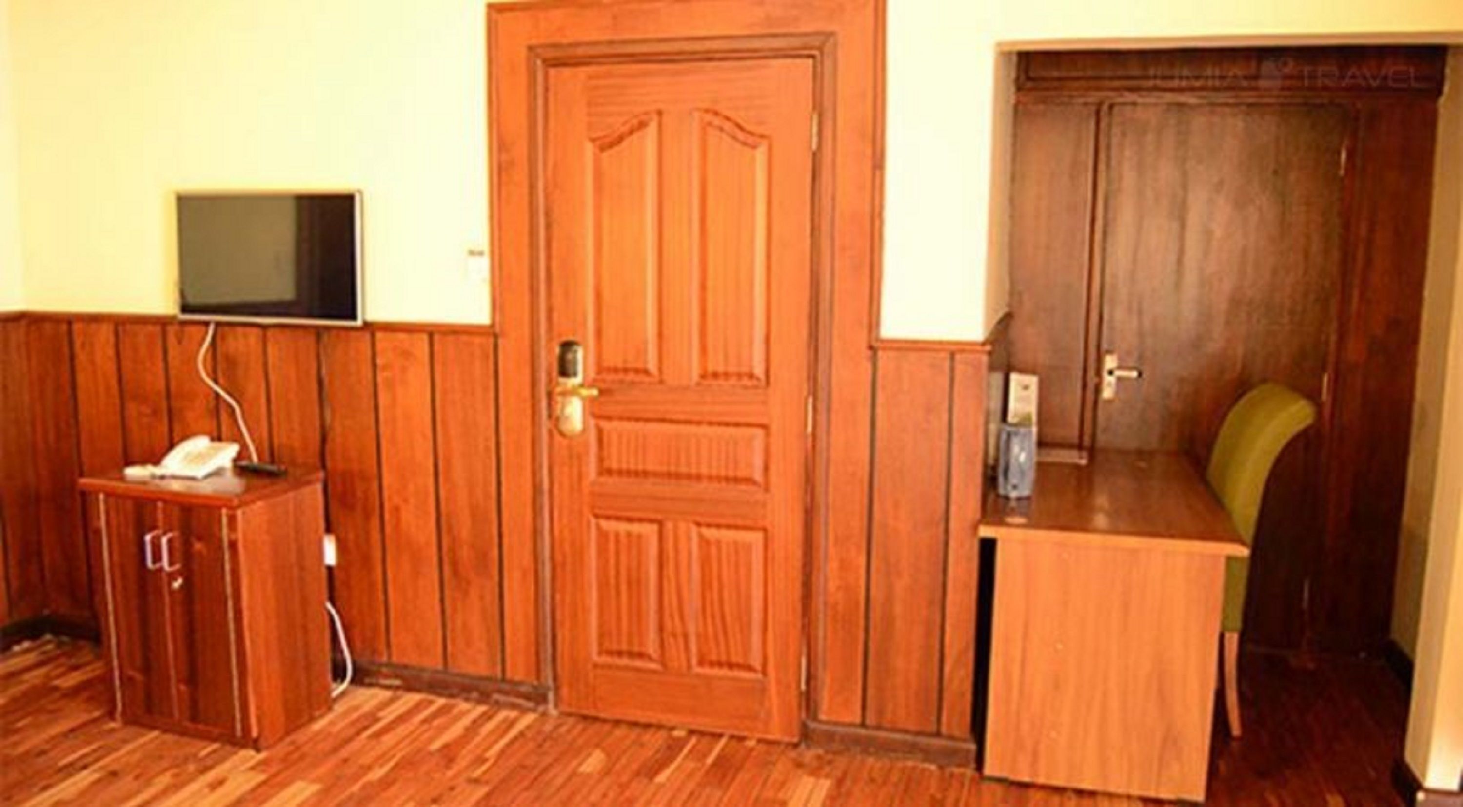 A room with a wooden door, a small desk with a chair, a television mounted on the wall, and a telephone on the desk.