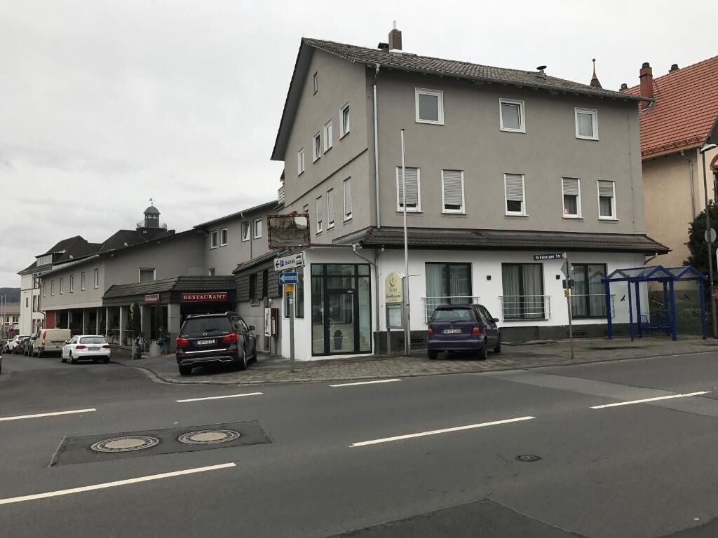 Un immeuble de deux étages avec un restaurant au rez-de-chaussée. Des voitures sont garées devant, et un arrêt de bus se trouve sur le côté droit.