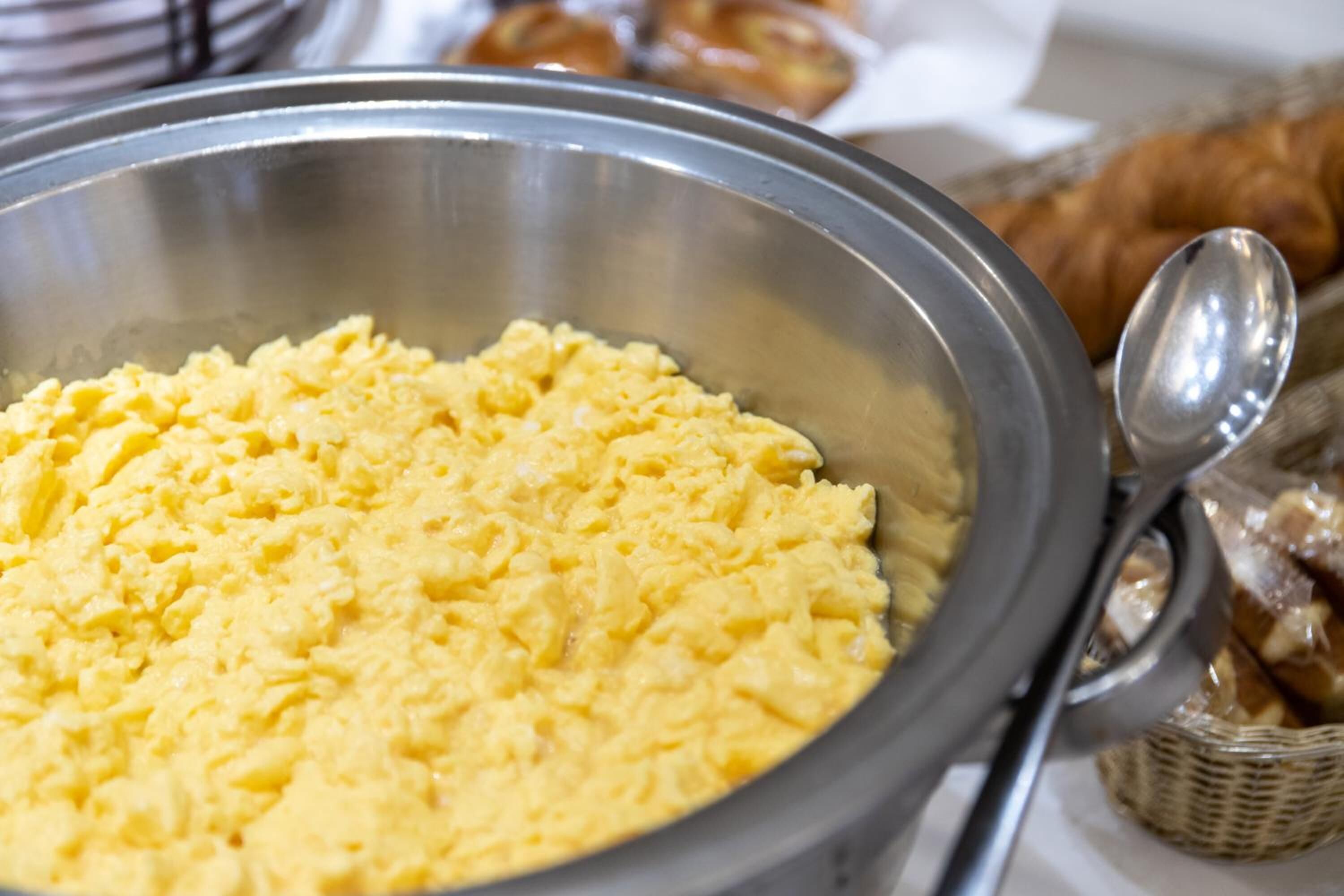 A stainless steel bowl filled with scrambled eggs, a spoon, and a basket of croissants.