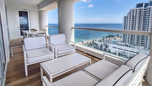 Een balkon met witte meubels, uitzicht op zee en een hoog gebouw op de achtergrond.